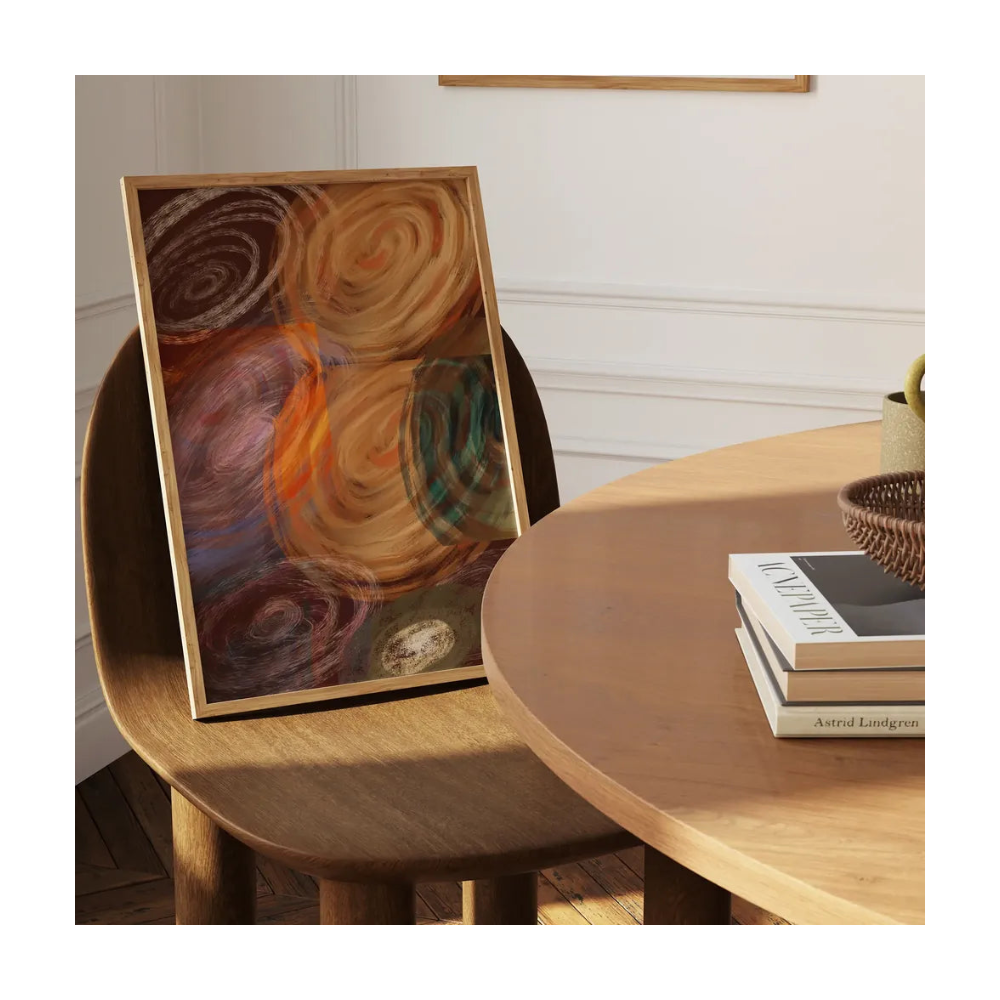 A framed abstract painting with swirling earthy tones of brown, orange, purple, and teal, leaning on a wooden chair beside a round wooden table with books.